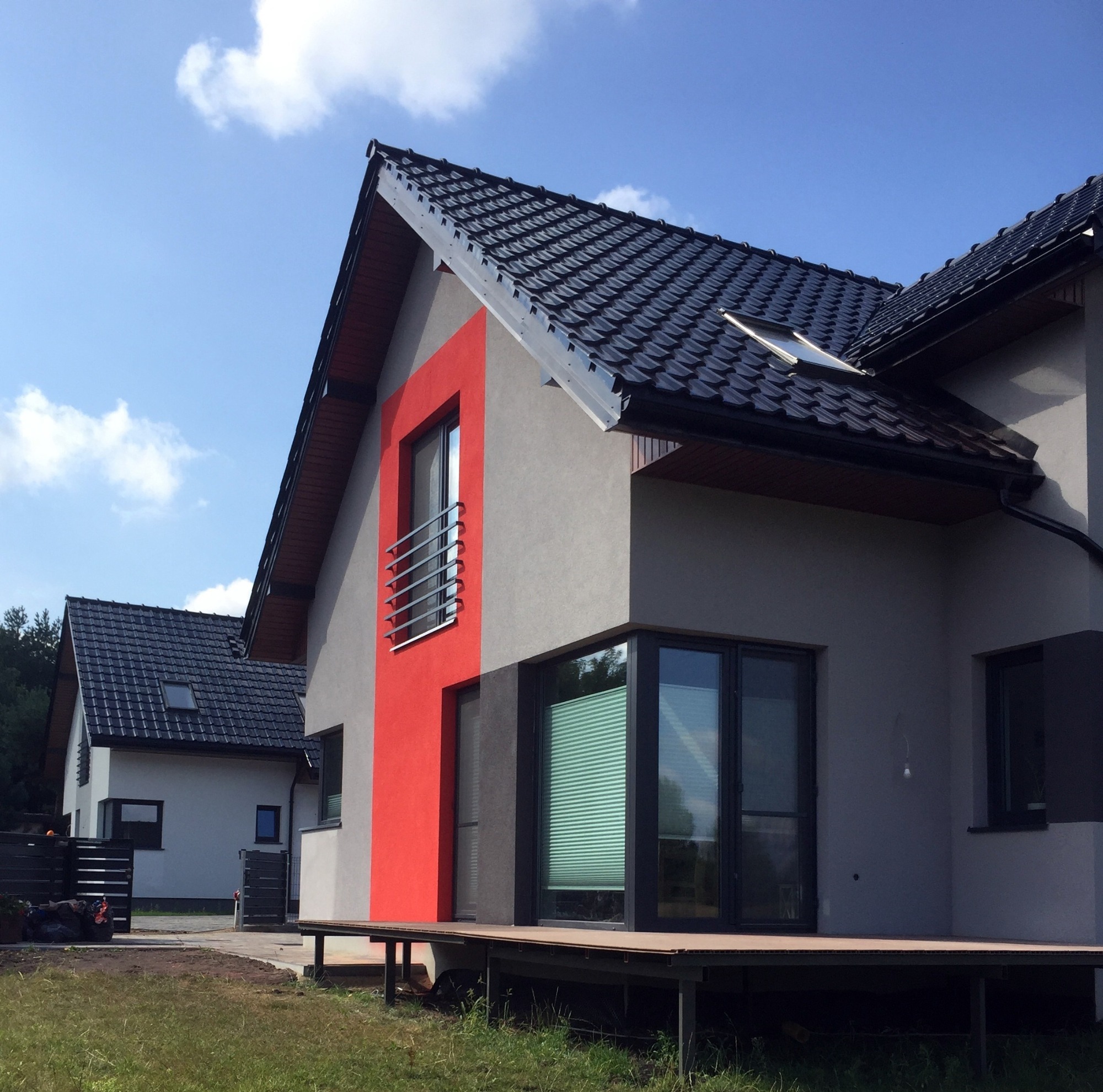 Nowoczesny dom z szarą elewacją i pionowym, czerwonym akcentem przy oknie z balkonem, czarny dach z dachówką ceramiczną i oknem dachowym.