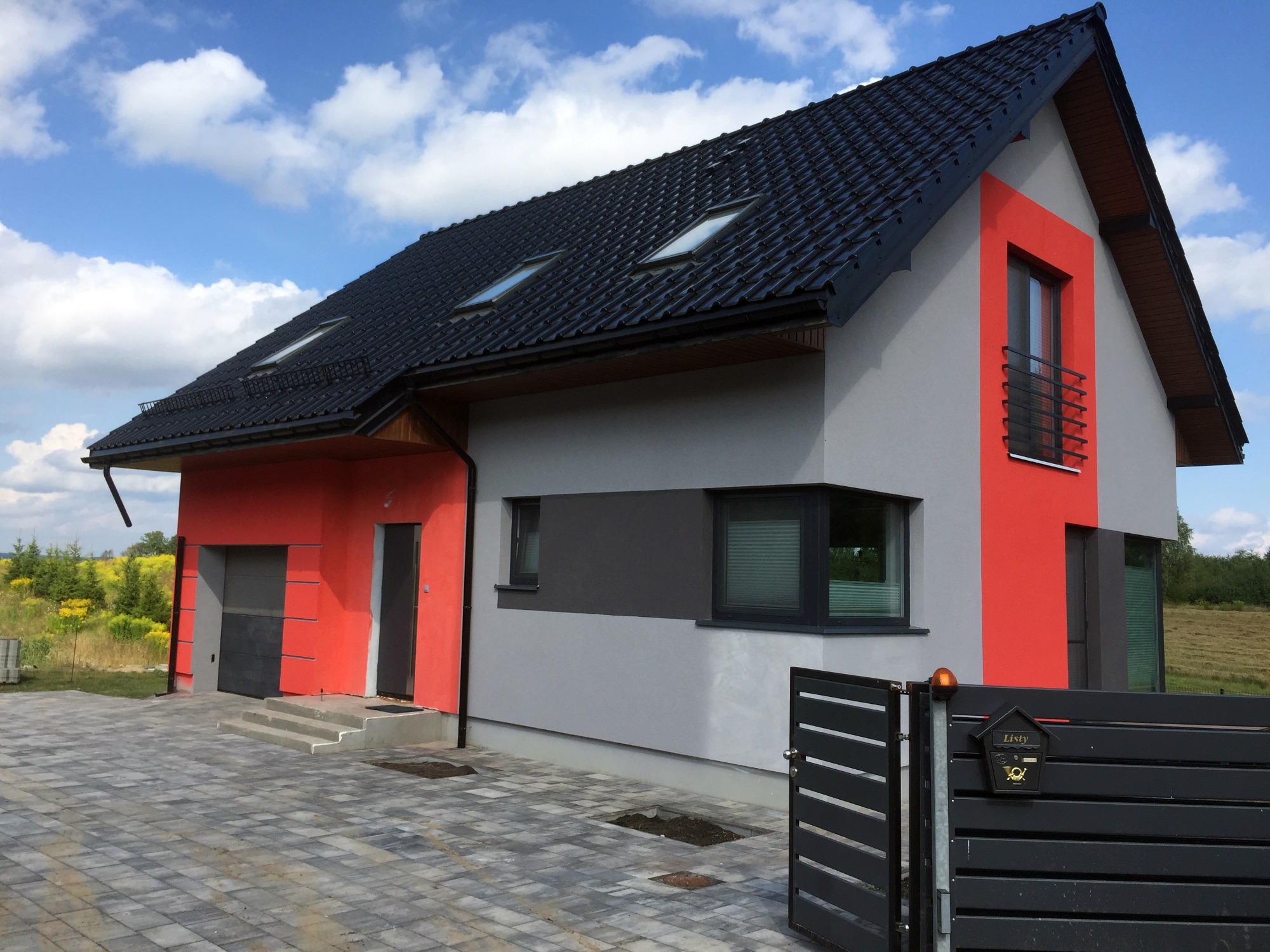 Nowoczesny dom jednorodzinny z szarą i czerwoną elewacją, czarnym dachem z oknami dachowymi, widok z zewnątrz na kostkę brukową i ogrodzenie z czarną skrzynką na listy.