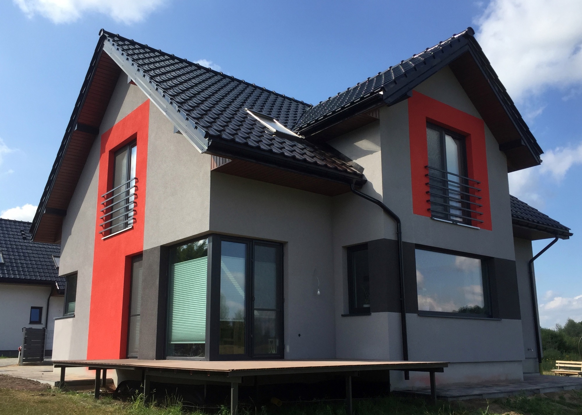 Nowoczesny dom jednorodzinny z szarą elewacją, akcentami w kolorze czerwonym wokół okien, czarnym dachem i drewnianym tarasem, widziany w słoneczny dzień.