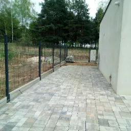 Nowoczesny taras z kostki brukowej w odcieniach szarości i beżu, przylegający do budynku z widocznym agregatem klimatyzacji, otoczony ogrodzeniem panelowym na betonowej podmurówce i zielenią.