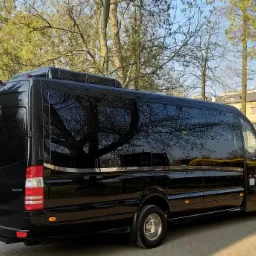 Czarny, luksusowy bus Mercedes Sprinter z przyciemnianymi szybami zaparkowany na brukowanej powierzchni, w tle drzewa i budynek z ceglanym kominem.