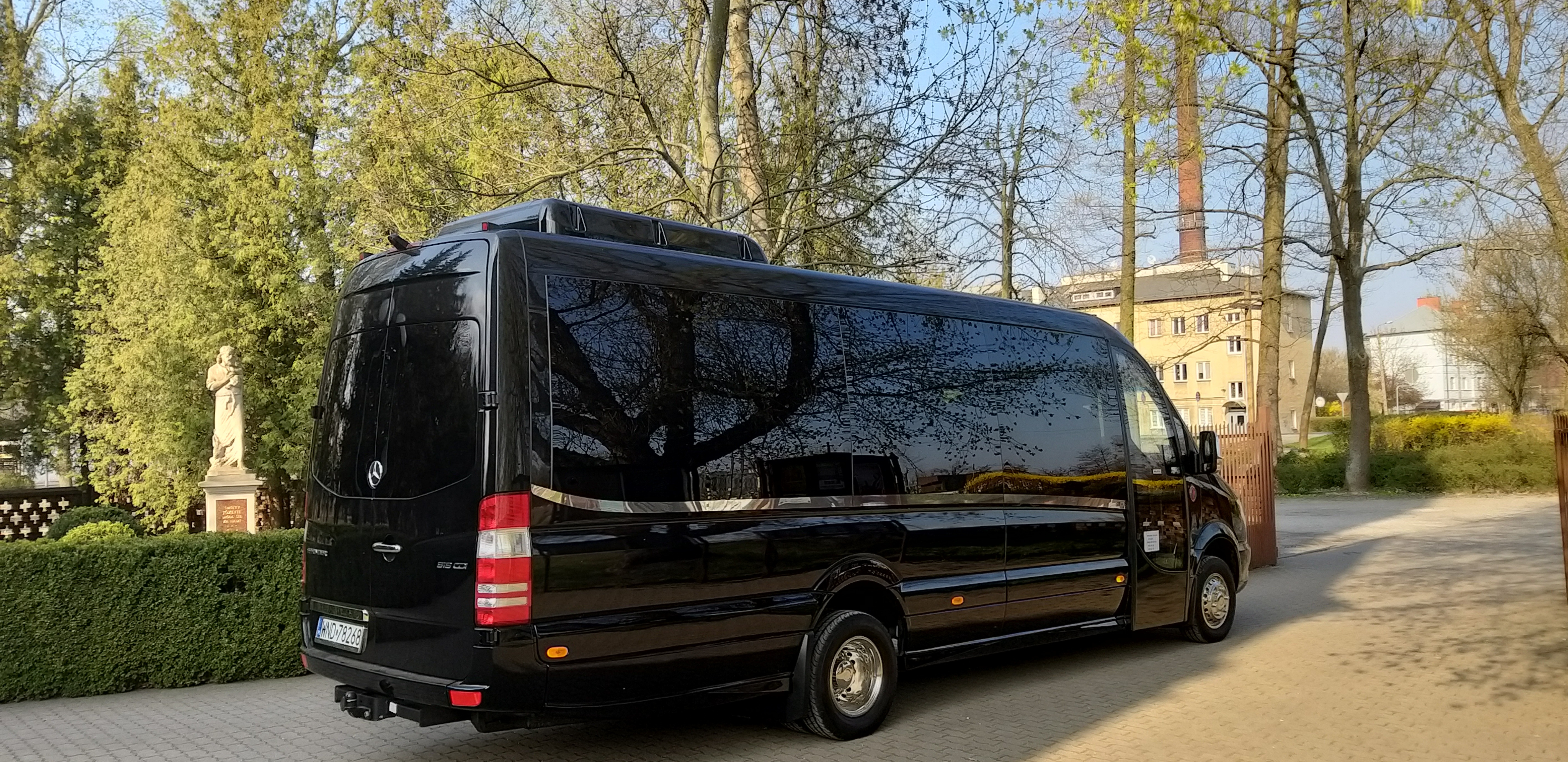 Czarny, luksusowy bus Mercedes Sprinter z przyciemnianymi szybami zaparkowany na brukowanej powierzchni, w tle drzewa i budynek z ceglanym kominem.