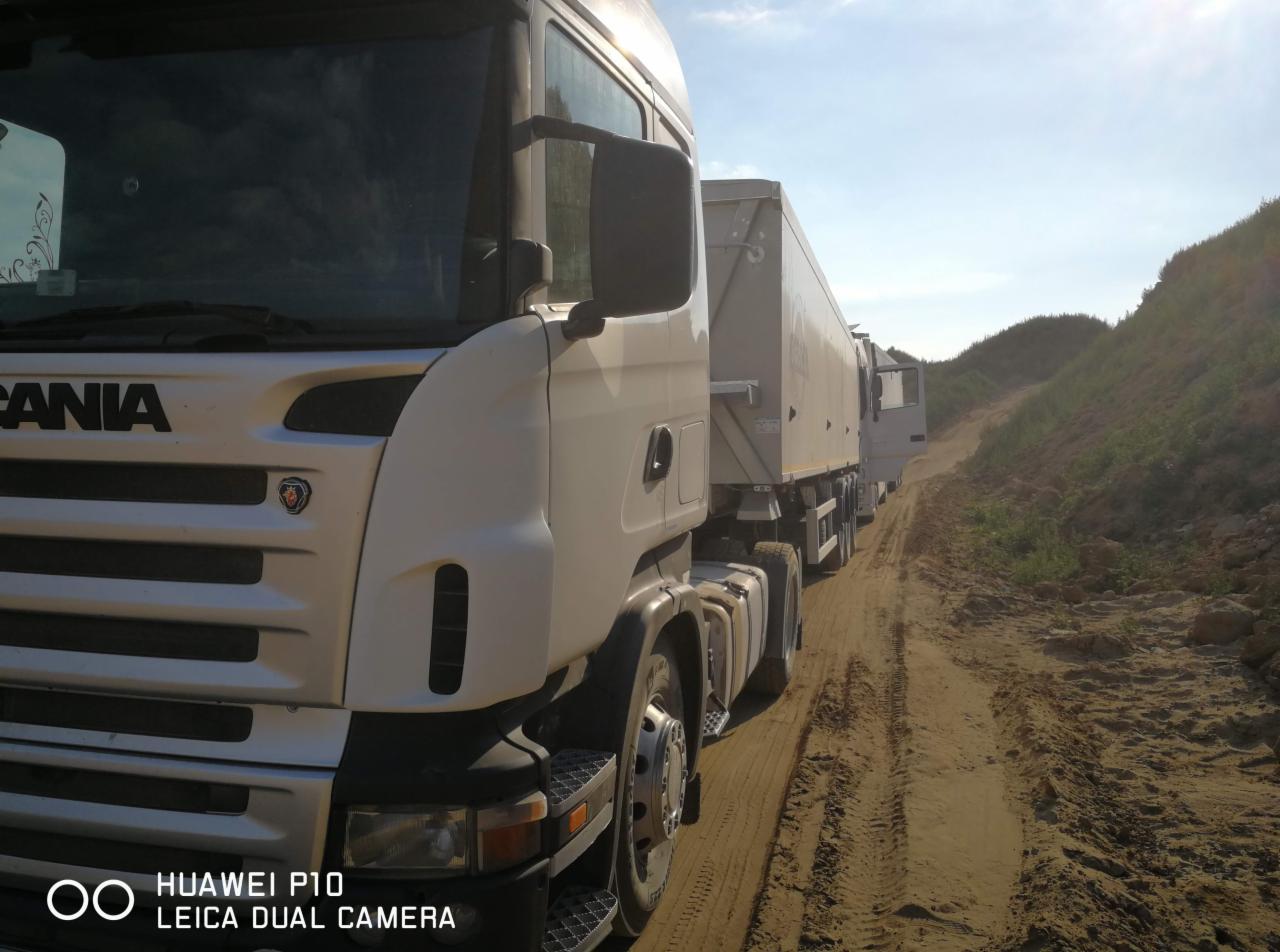 Kolumna białych ciężarówek marki Scania z naczepami skrzyniowymi, jadąca po piaszczystej drodze w terenie pagórkowatym, w słoneczny dzień.