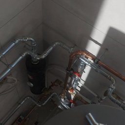IHR - SYSTEMS - Złożona instalacja hydrauliczna z rur miedzianych i stalowych w pomieszczeniu wyłożonym białymi płytkami, widok z dołu, z elementami zbiornika i przewodami elektrycznymi.
