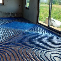 IHR-SYSTEMS PIOTR GĄDEK - Niebieskie rury ogrzewania podłogowego ułożone na folii w surowym wnętrzu domu z widokiem na zewnątrz przez okno. Widoczna drabina i drewniana konstrukcja dachu.