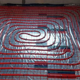 IHR-SYSTEMS PIOTR GĄDEK - Czerwone rury ogrzewania podłogowego ułożone na srebrnej folii z nadrukiem na podłożu w surowym pomieszczeniu. Widok z góry na spiralny układ rur.
