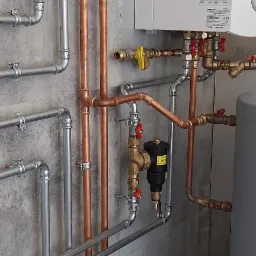 Nowa instalacja grzewcza z miedzianymi i stalowymi rurami, zaworami i pompami obiegowymi na tle betonowej ściany. Widoczny zasobnik ciepłej wody i fragment kotła.