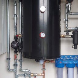 IHR - SYSTEMS - Czarny zbiornik Viqtis z manometrami, otoczony instalacją hydrauliczną z rur miedzianych i stalowych, filtry wody z niebieskimi obudowami, biała szafka na dole.