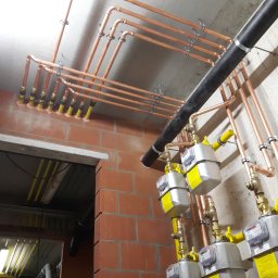 IHR - SYSTEMS - Miedziana instalacja gazowa z licznikami, zamontowana przy ceglanej ścianie i betonowym suficie. Widoczne żółte zawory i czarny przewód wentylacyjny.