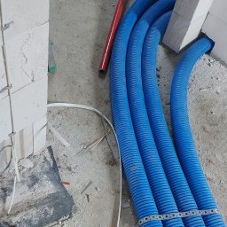 IHR-SYSTEMS PIOTR GĄDEK - Niebieskie rury karbowane, częściowo zamocowane, prowadzone wzdłuż ściany w surowym wnętrzu. Czerwona rura i narzędzia w tle. Instalacja ogrzewania podłogowego w trakcie.