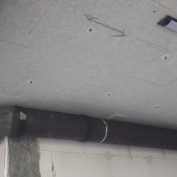 IHR-SYSTEMS PIOTR GĄDEK - Czarne, zaizolowane rury wentylacyjne przymocowane do sufitu z płyt. Widoczne mocowania, strzałki na suficie i lampa. Instalacja w surowym wnętrzu.