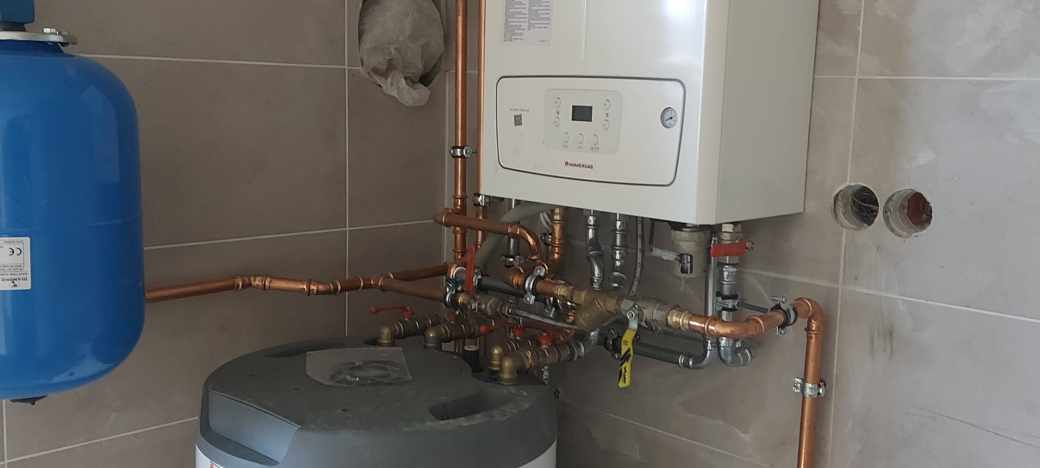 Biały wiszący piec gazowy Immergas z miedzianą instalacją, niebieski zbiornik wyrównawczy i szary zasobnik ciepłej wody w kotłowni wykończonej płytkami ceramicznymi.