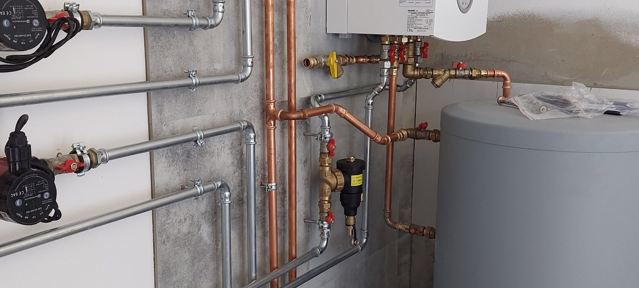 Nowa instalacja grzewcza z miedzianymi i stalowymi rurami, zaworami i pompami obiegowymi na tle betonowej ściany. Widoczny zasobnik ciepłej wody i fragment kotła.