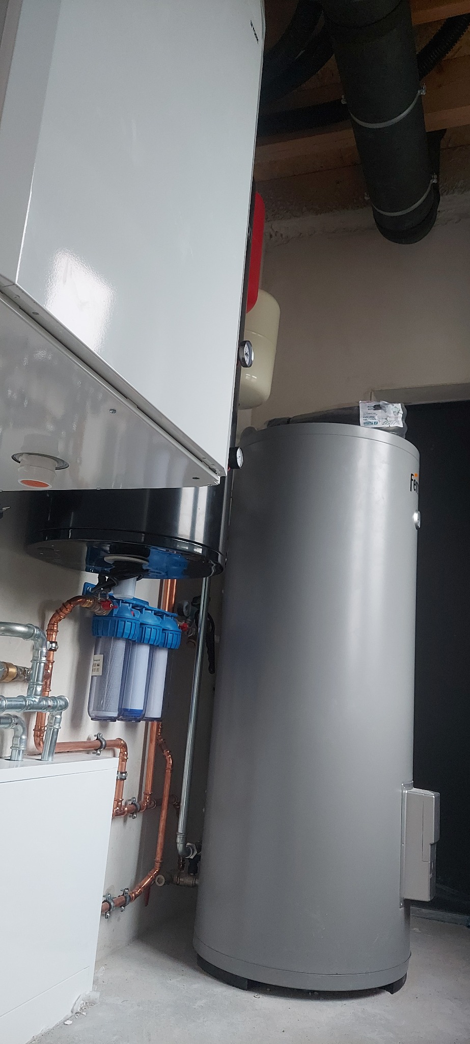 Nowoczesny system grzewczy z białym piecem, szarym zasobnikiem i filtrami wody na miedzianych rurach. Widoczne elementy instalacji pod sufitem.