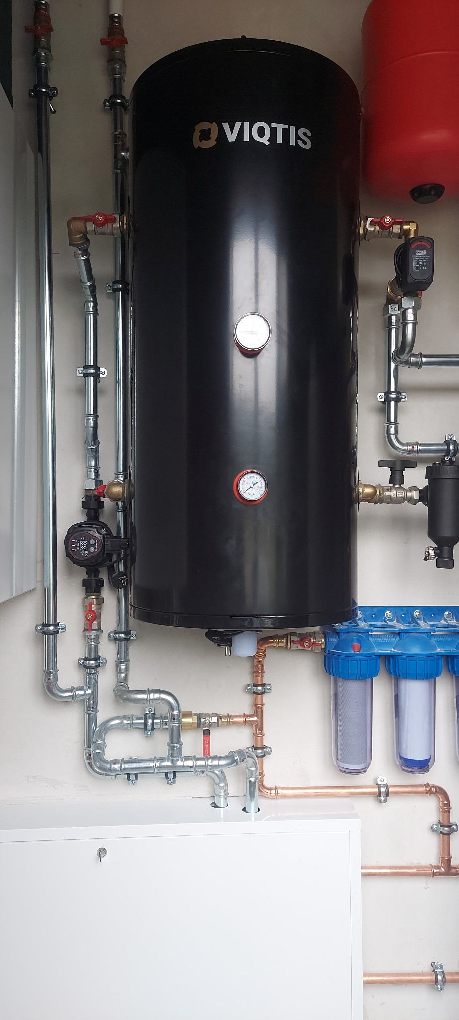 Czarny zbiornik Viqtis z manometrami, otoczony instalacją hydrauliczną z rur miedzianych i stalowych, filtry wody z niebieskimi obudowami, biała szafka na dole.