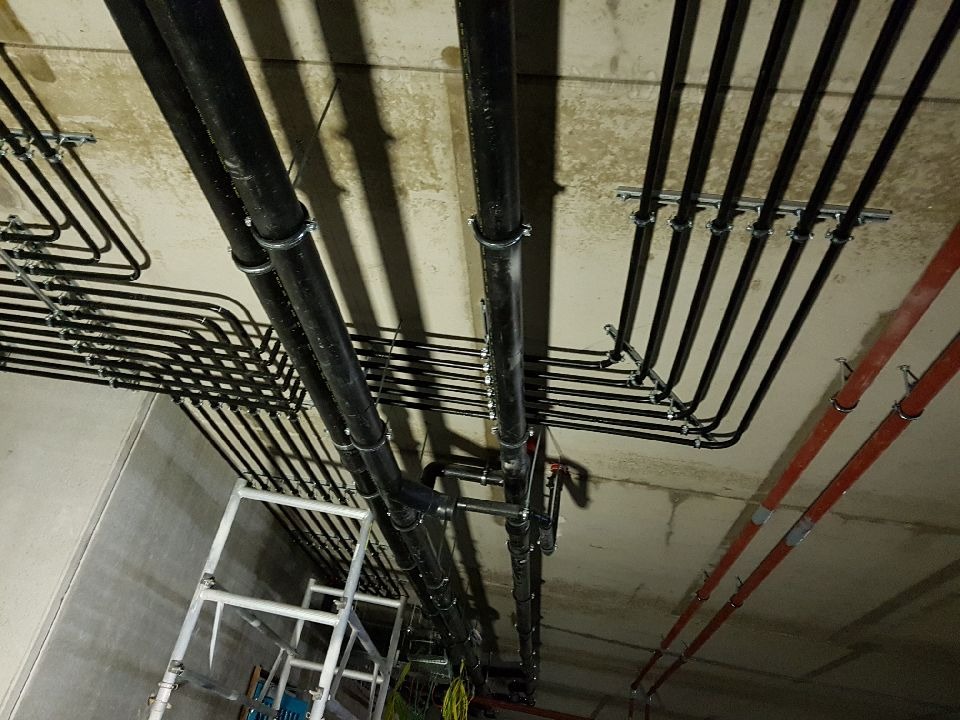 Czarny system rur pod sufitem, wsparty na metalowych uchwytach, z widoczną drabiną i fragmentami instalacji w tle. Ujęcie z dołu, podkreślające złożoność instalacji.