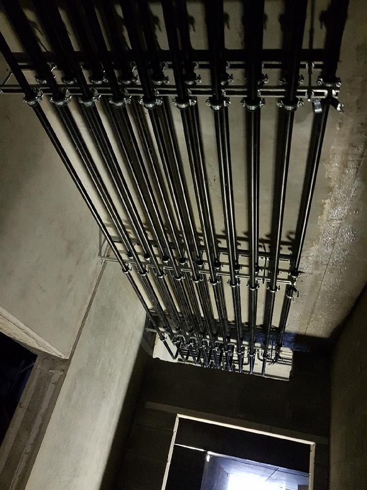 Równolegle ułożone, czarne rury instalacyjne pod sufitem, mocowane metalowymi uchwytami. Widok z dołu, surowy beton i fragment otworu drzwiowego w tle.