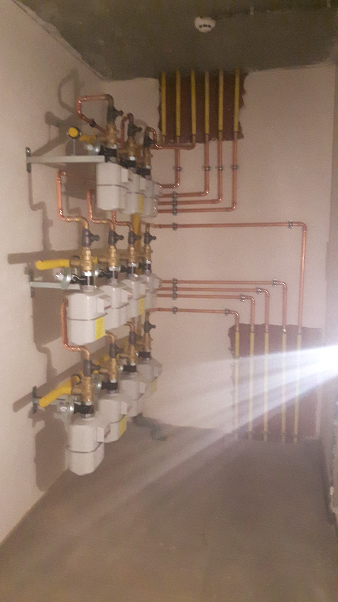 Rząd liczników gazowych z miedzianymi rurami, zamontowanych na ścianie w surowym wnętrzu. Widoczne pionowe rury w żółtej izolacji.