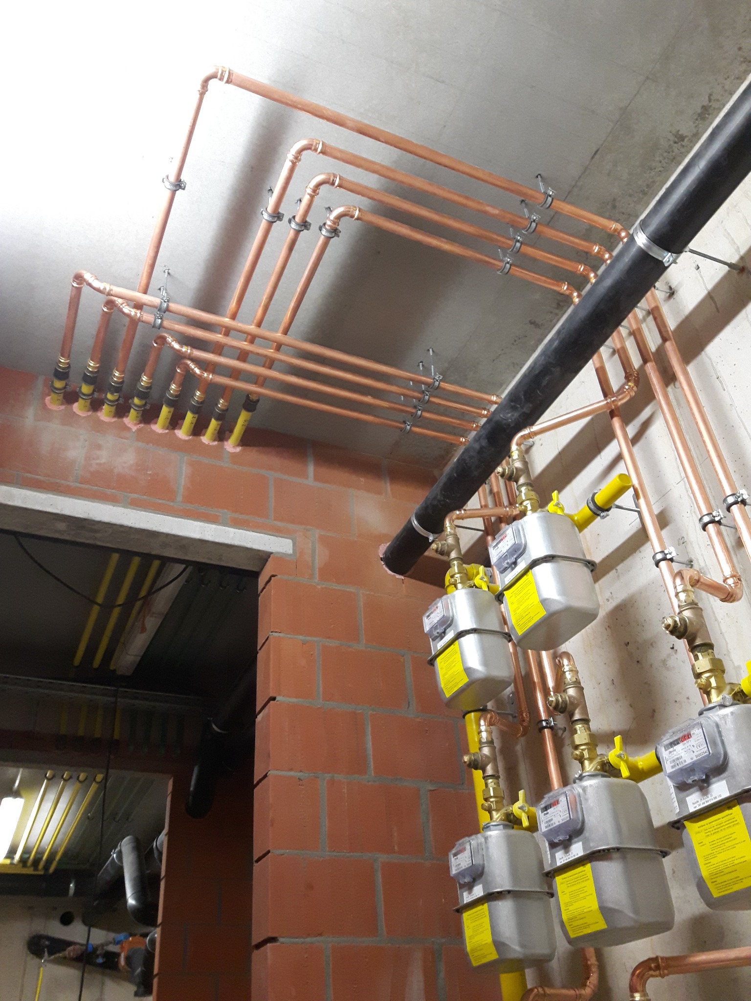 Miedziana instalacja gazowa z licznikami, zamontowana przy ceglanej ścianie i betonowym suficie. Widoczne żółte zawory i czarny przewód wentylacyjny.