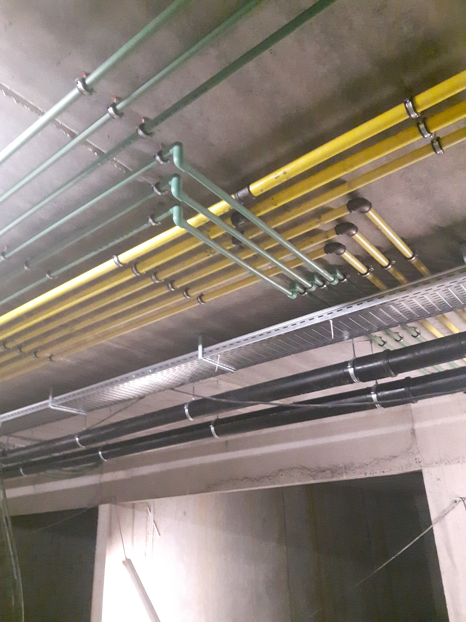 Różnokolorowe rury instalacyjne (zielone, żółte, czarne) pod sufitem, zamocowane metalowymi obejmami. Instalacje w budynku w Gdańsku.