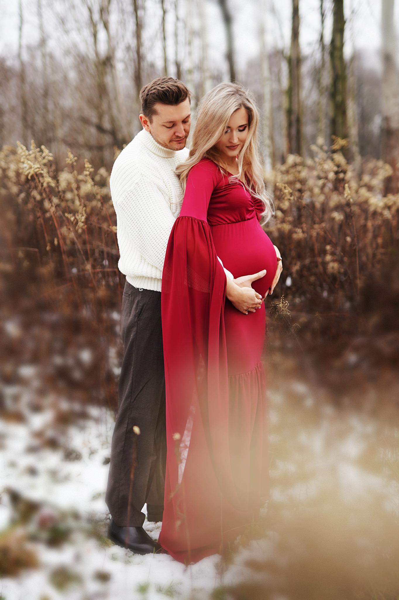 Uśmiechnięta, ciężarna blondynka w długiej, czerwonej sukni z rozszerzanymi rękawami, obejmowana przez partnera w białym swetrze na tle zimowego krajobrazu z resztkami śniegu.