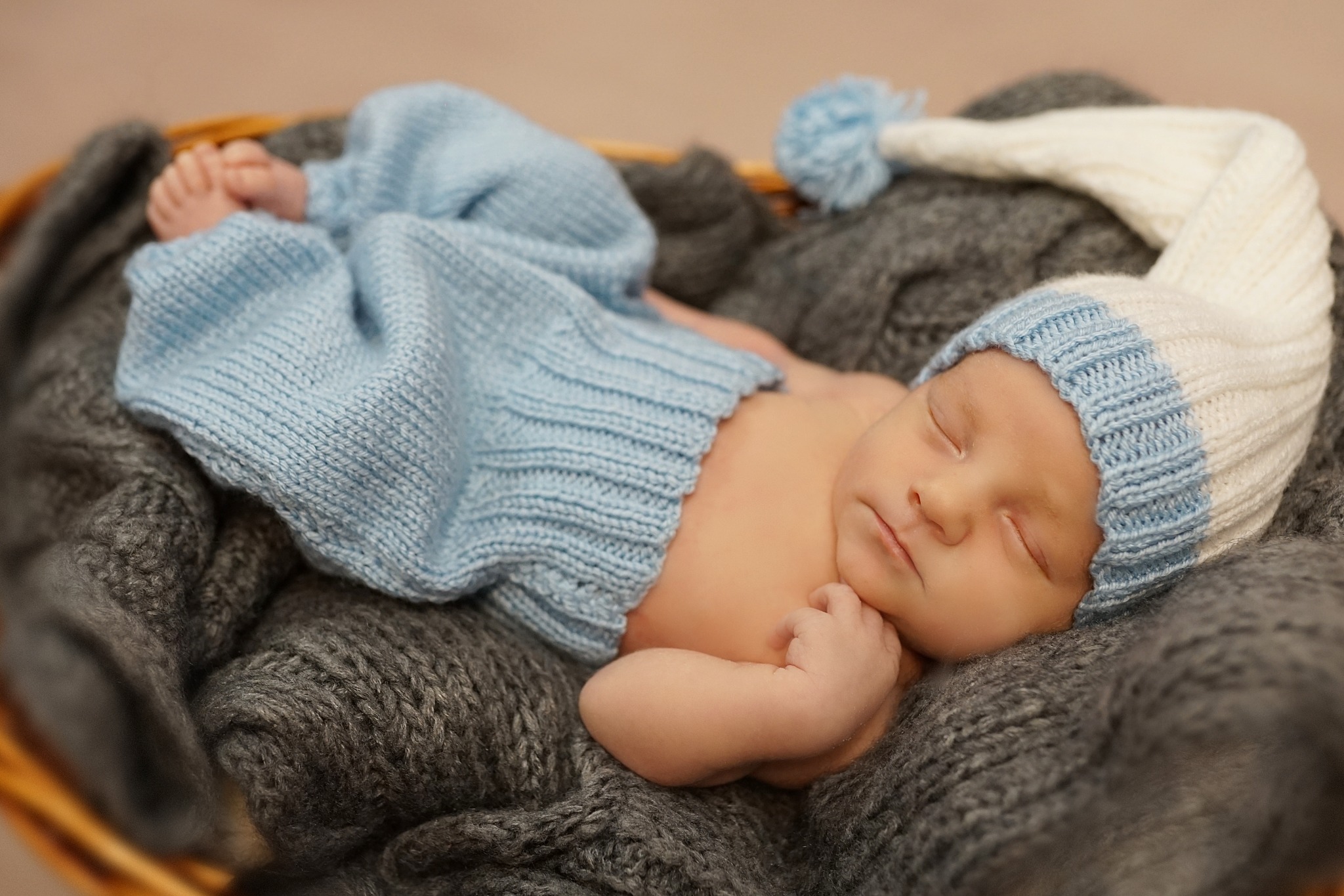 Śpiący noworodek w niebieskim, dzierganym ubranku i czapce z pomponem, ułożony w wiklinowym koszu na szarym, dzierganym kocyku.