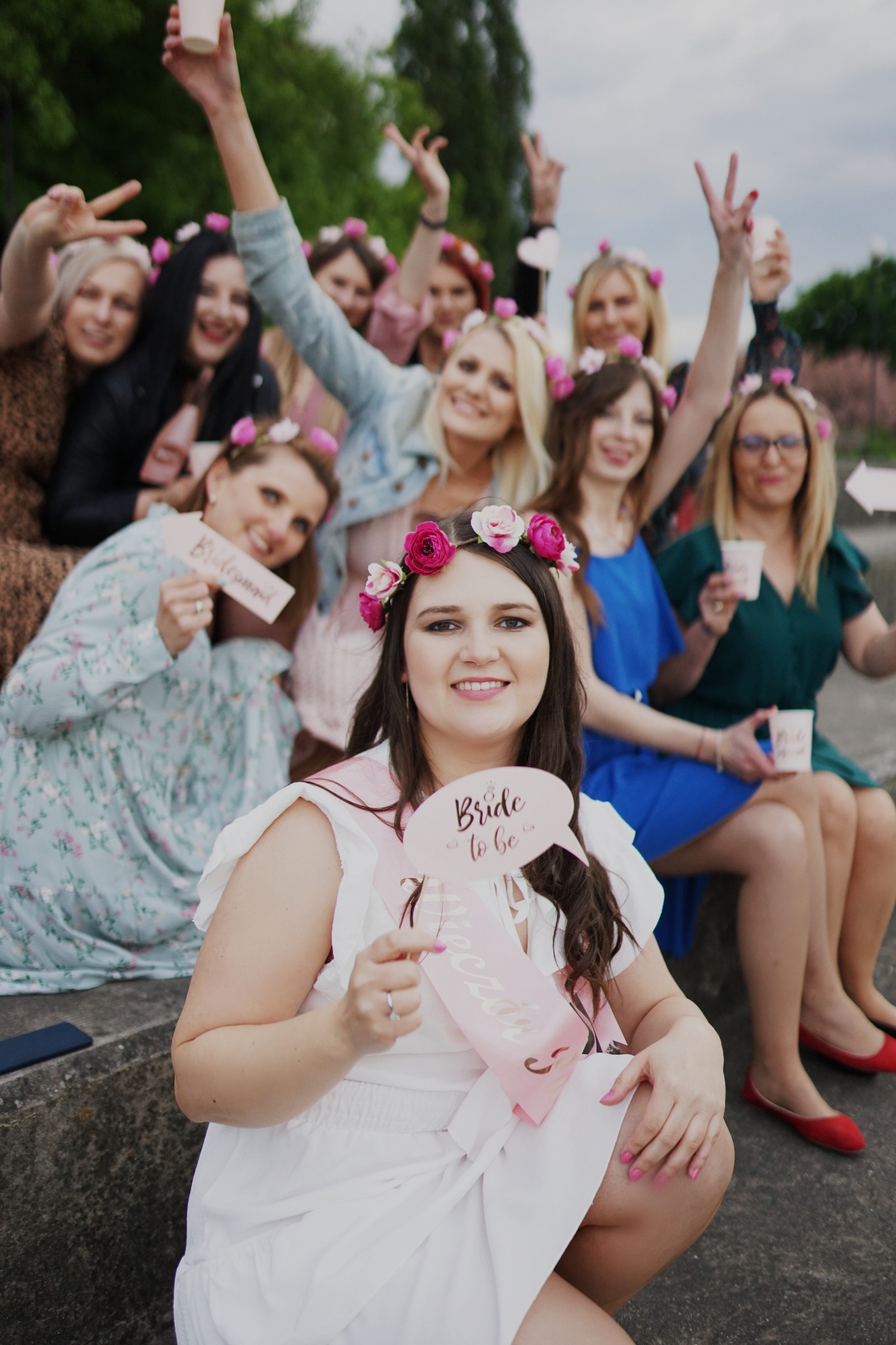 Panna młoda w białej sukience i z szarfą 'Wieczór Panieński' trzymająca rekwizyt z napisem 'Bride to be', otoczona uśmiechniętymi kobietami w wiankach z kwiatów podczas wieczoru panieńskiego.