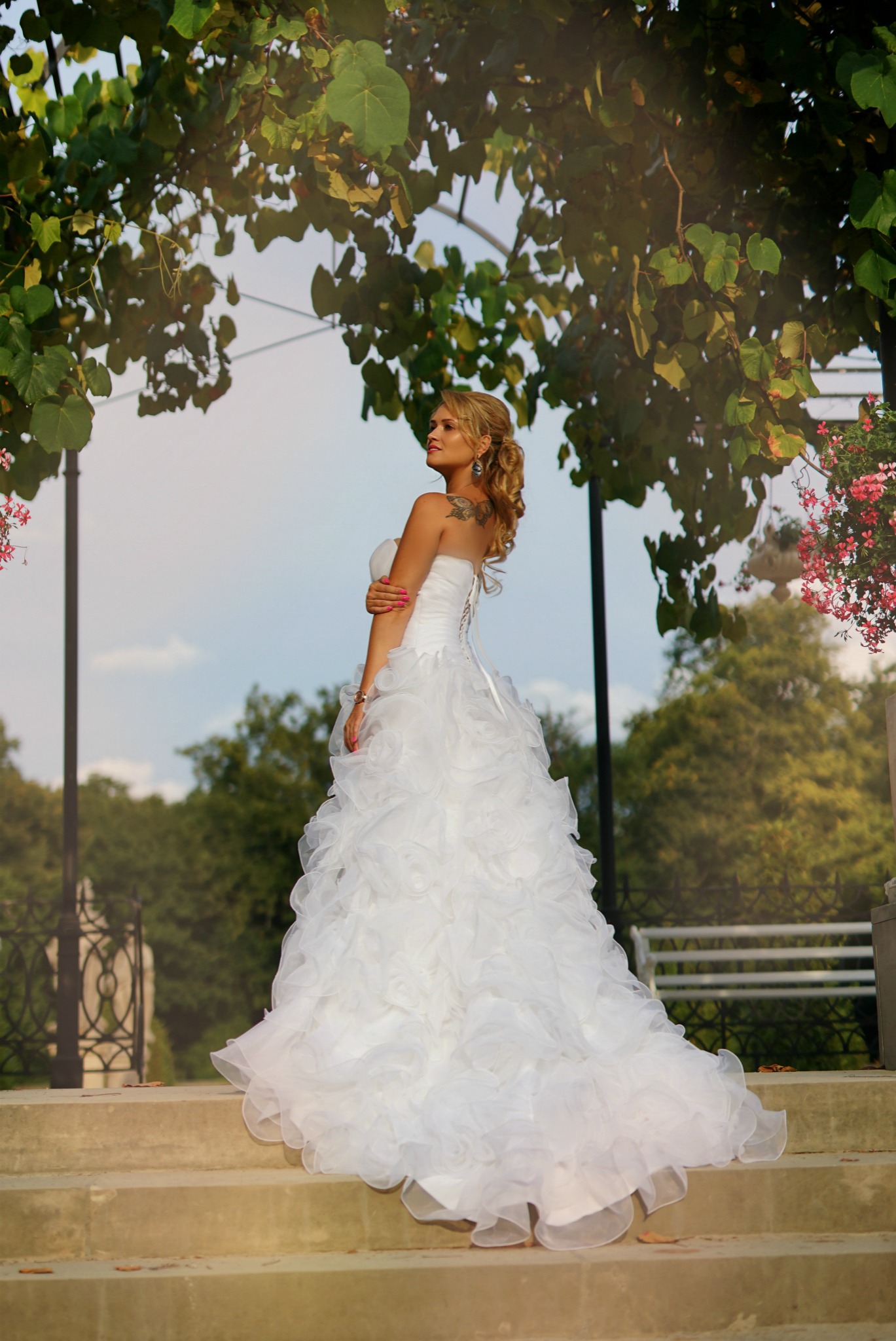 Panna młoda w białej sukni z falbanami, stojąca na schodach pod altaną porośniętą zielenią, z tatuażem na ramieniu i upiętymi blond włosami, spogląda w bok.