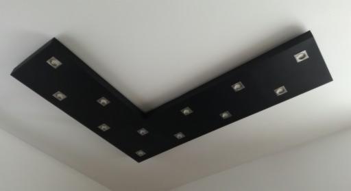 Czarny, podwieszany sufit z wbudowanymi, kwadratowymi lampkami LED, tworzący kąt prosty na białym suficie w jasnym pomieszczeniu.