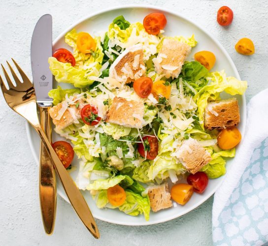 Sałatka Cezar z pomidorkami koktajlowymi, grzankami i parmezanem na talerzu z złotymi sztućcami i lnianą ściereczką.