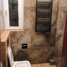 Wykończona łazienka z brązowymi marmurkowymi płytkami, białym sedesem, grzejnikiem drabinkowym i drewnianą podłogą, widoczne niedociągnięcia w postaci wystających kabli.