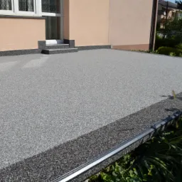 Szara, drobnoziarnista nawierzchnia tarasu z żywicy, z ciemniejszym obramowaniem, przylegająca do ściany budynku z oknem i schodkami, otoczona zielenią.