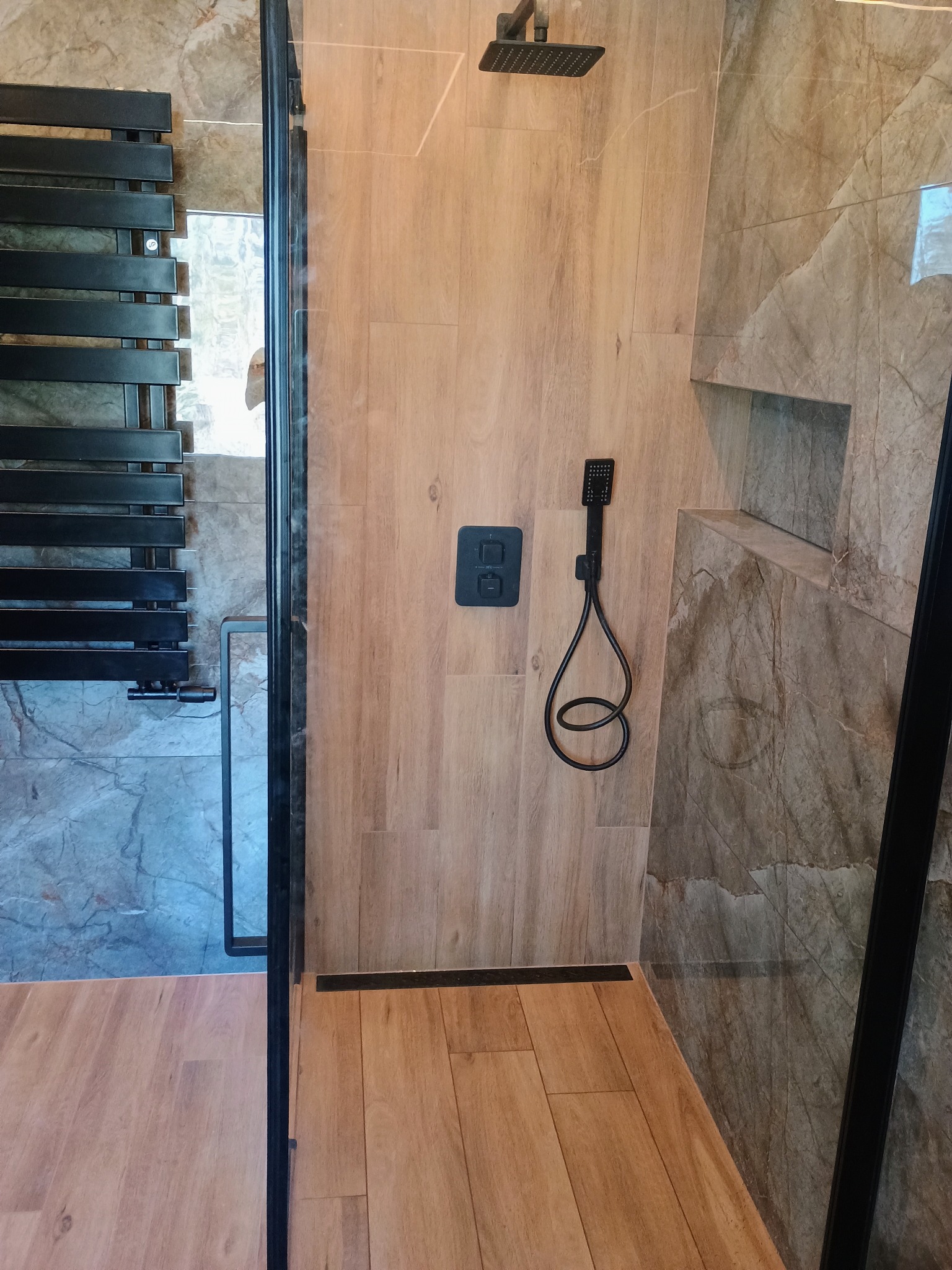 Nowoczesna kabina prysznicowa z panelami imitującymi drewno, czarną armaturą i grzejnikiem drabinkowym, widok z perspektywy wejścia.