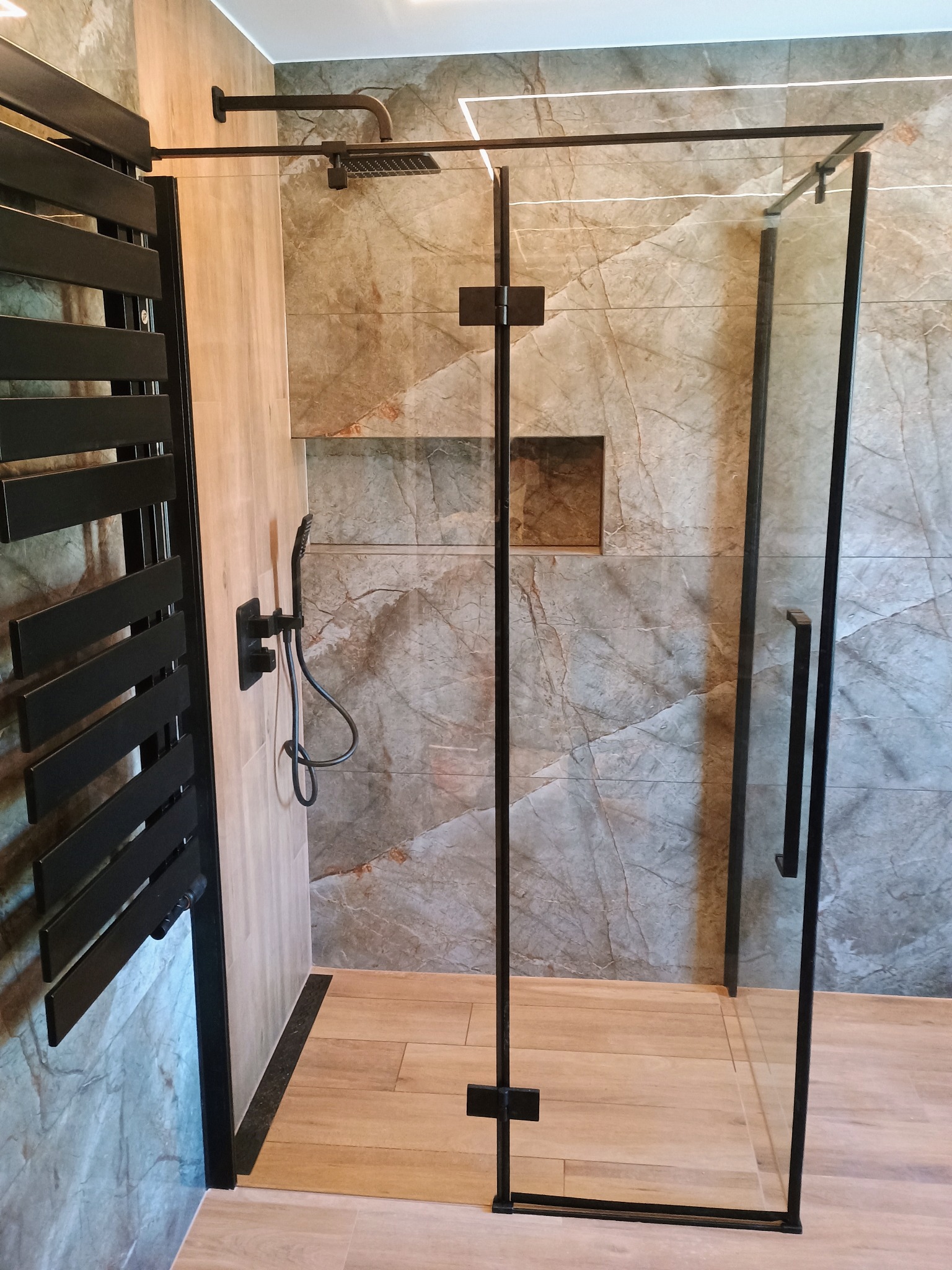 Nowoczesna kabina prysznicowa z czarnymi profilami i deszczownicą zamontowana w wykończonej łazience z płytkami imitującymi marmur i drewnopodobną podłogą.