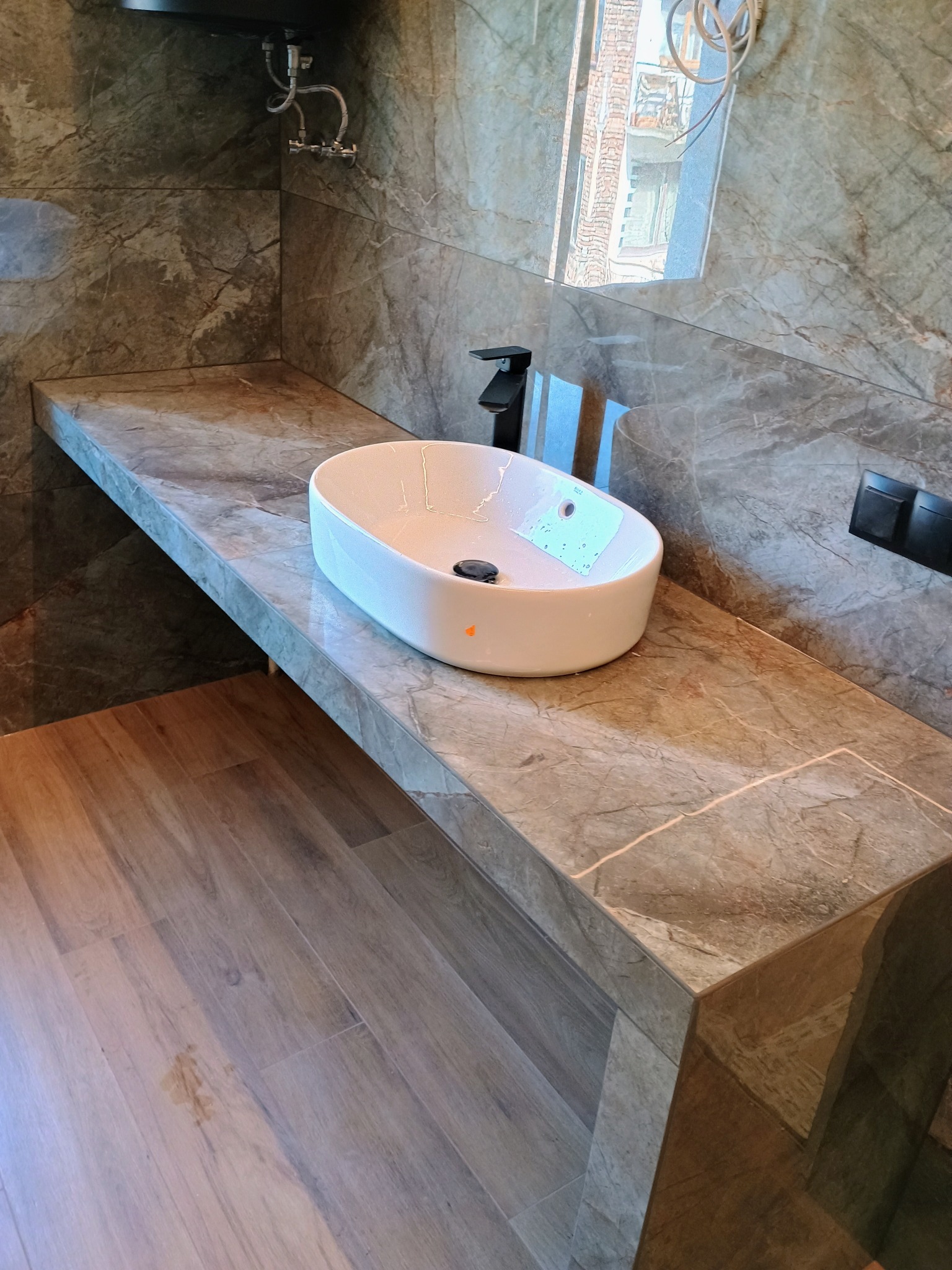 Nowoczesna łazienka z umywalką nablatową w kształcie elipsy, czarną baterią i marmurową zabudową, odbicie okna w lustrze, podłoga drewnopodobna.