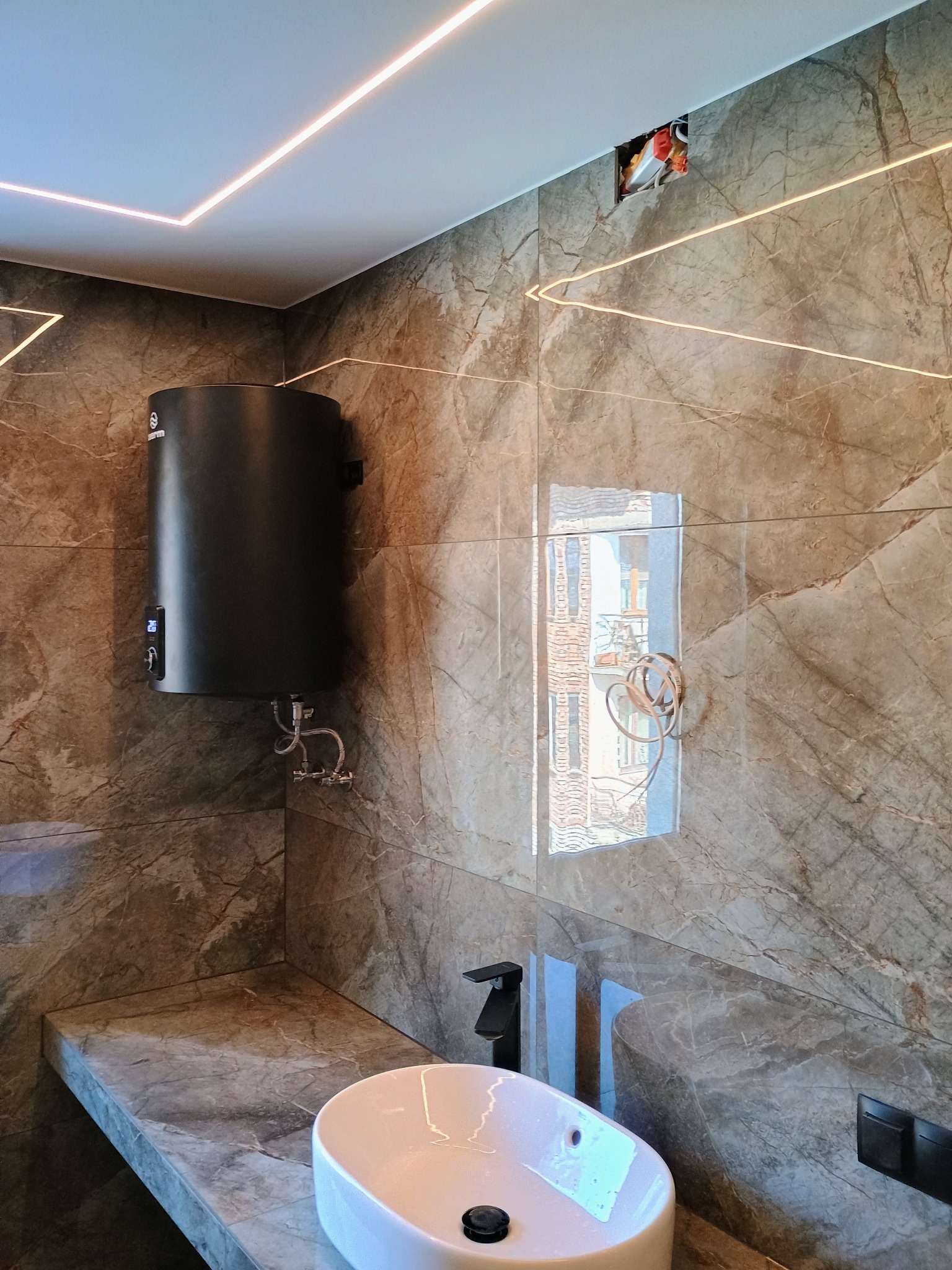Nowoczesna łazienka z marmurowymi płytkami, podświetlanym sufitem LED, owalną umywalką nablatową i czarnym bojlerem elektrycznym na ścianie. Widoczne niedokończone podłączenie oświetlenia w suficie.