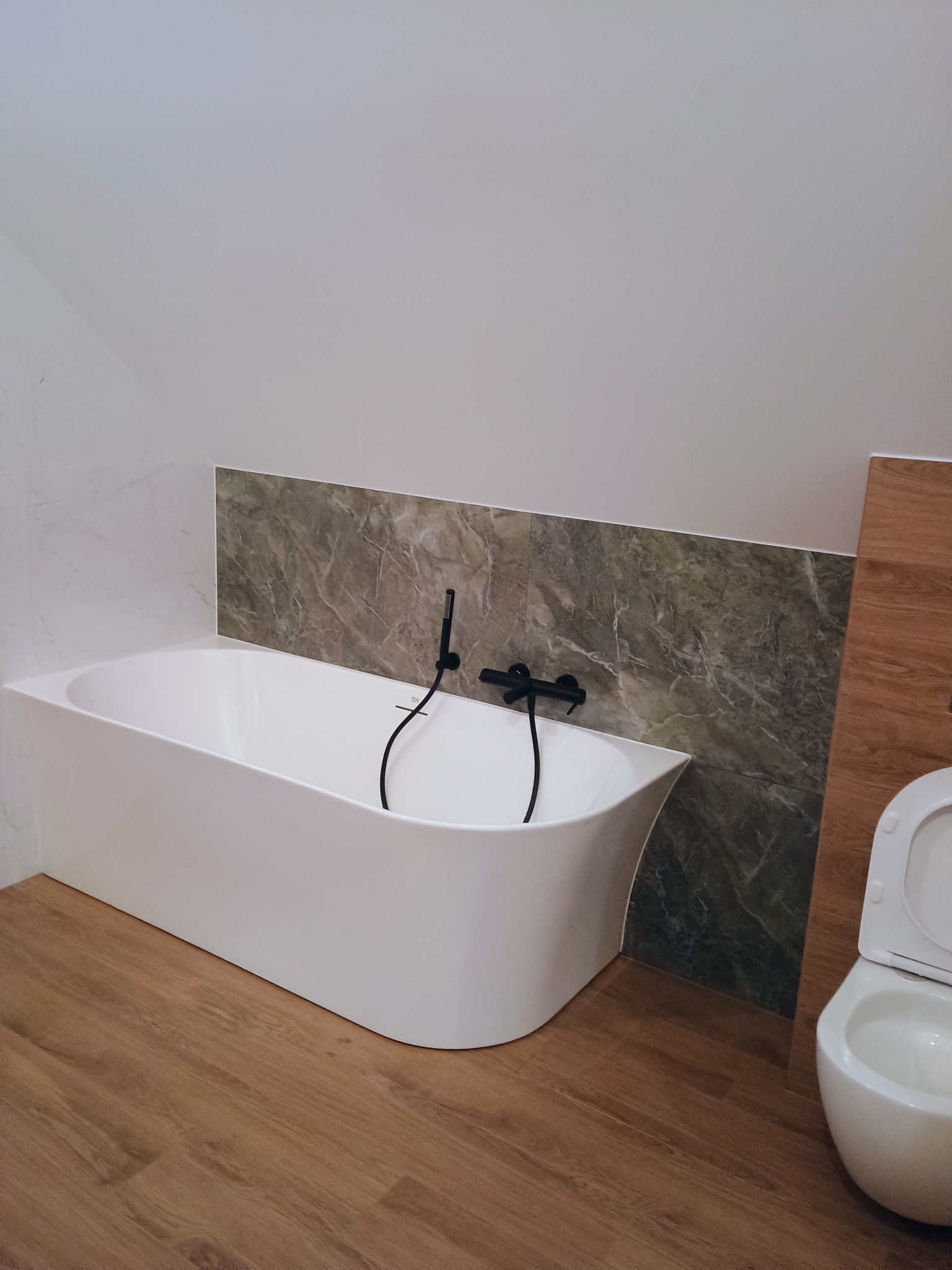 Nowoczesna biała wanna wolnostojąca z czarną armaturą na tle marmurowej ściany w łazience na poddaszu z drewnopodobną podłogą i częściowo widoczną toaletą.