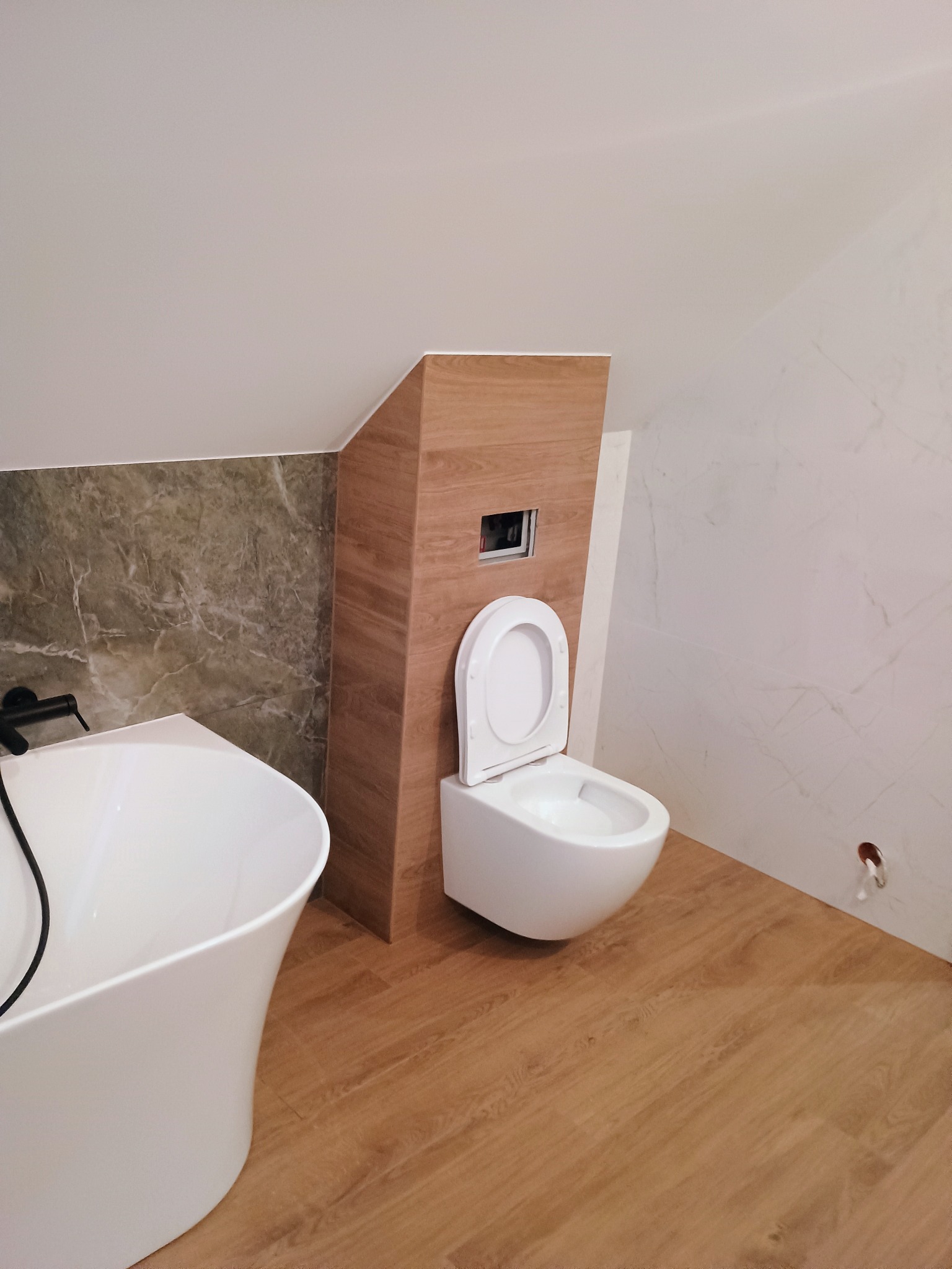 Nowoczesna łazienka na poddaszu ze skośnym sufitem, z wolnostojącą białą wanną, podwieszaną toaletą z drewnianą obudową stelaża, marmurowymi płytkami i drewnopodobną podłogą.