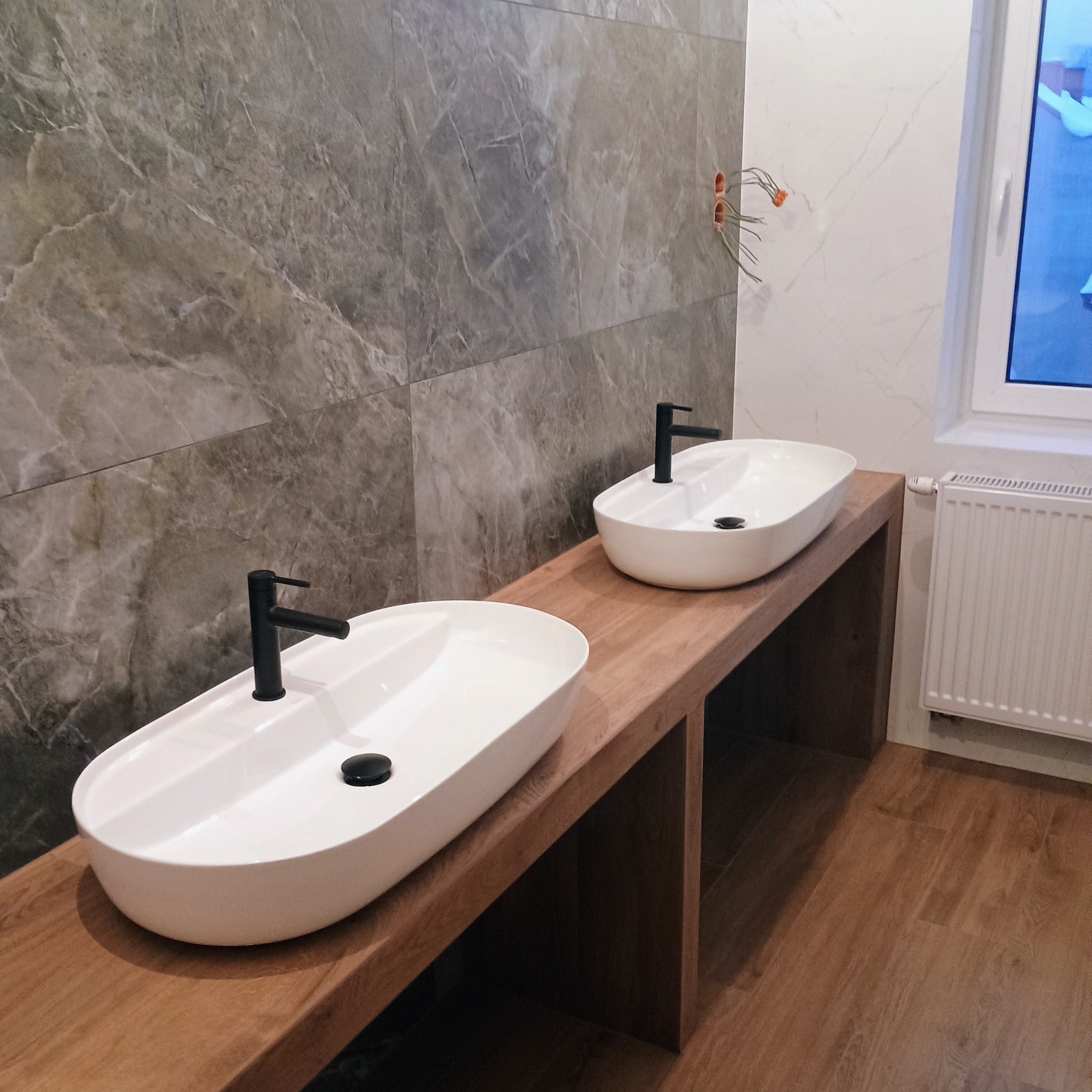 Dwie białe, owalne umywalki nablatowe z czarną armaturą, osadzone na drewnianym blacie w łazience wykończonej szarymi płytkami imitującymi marmur. Widoczne wystające kable elektryczne oraz biały...