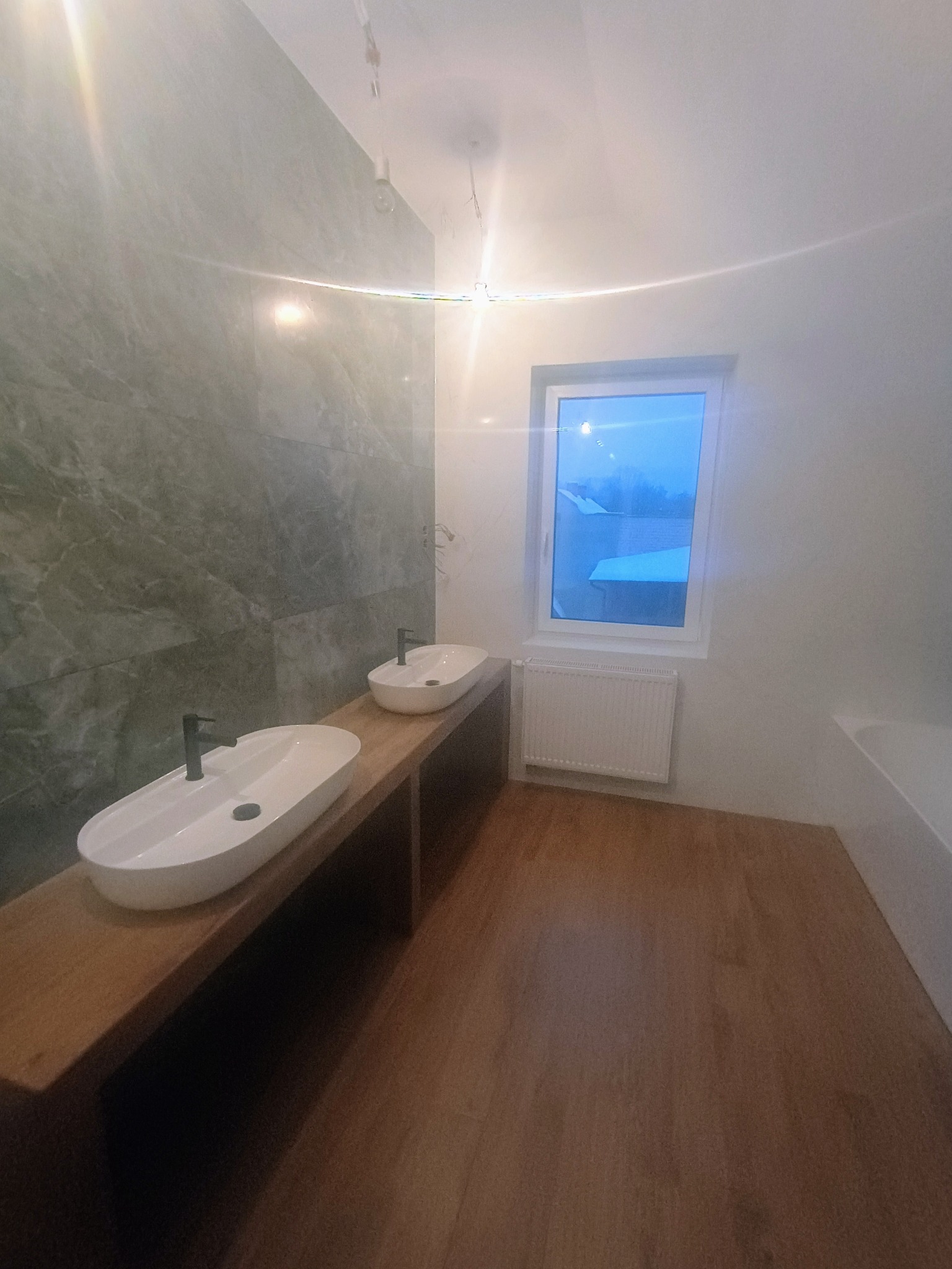 Nowoczesna łazienka z dwoma owalnymi umywalkami na drewnianym blacie, marmurowe płytki na ścianie, widok na zewnątrz przez okno.