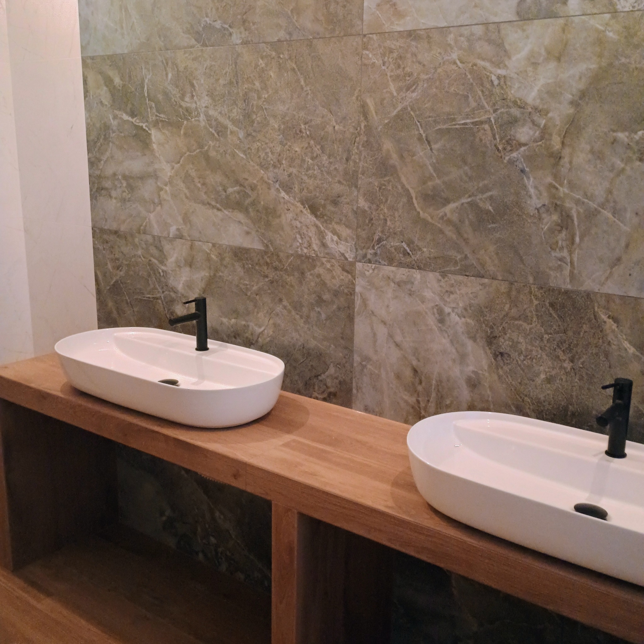 Dwie białe umywalki nablatowe o owalnym kształcie z czarnymi bateriami, zamontowane na drewnianym blacie z półkami, na tle ściany wyłożonej płytkami imitującymi marmur.