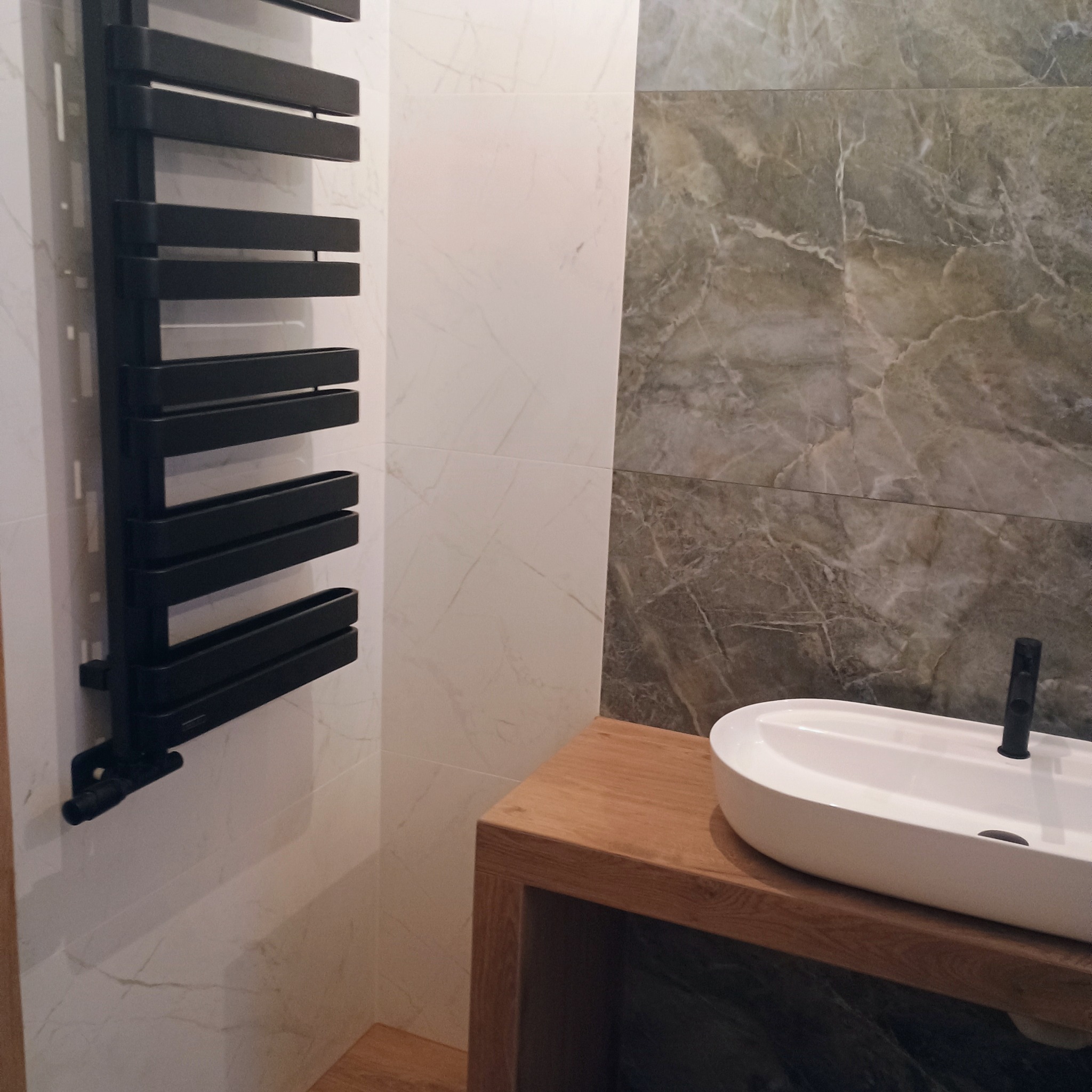 Wykończona łazienka z owalną umywalką nablatową na drewnianym blacie, czarnym grzejnikiem drabinkowym i płytkami imitującymi marmur w odcieniach szarości i bieli.