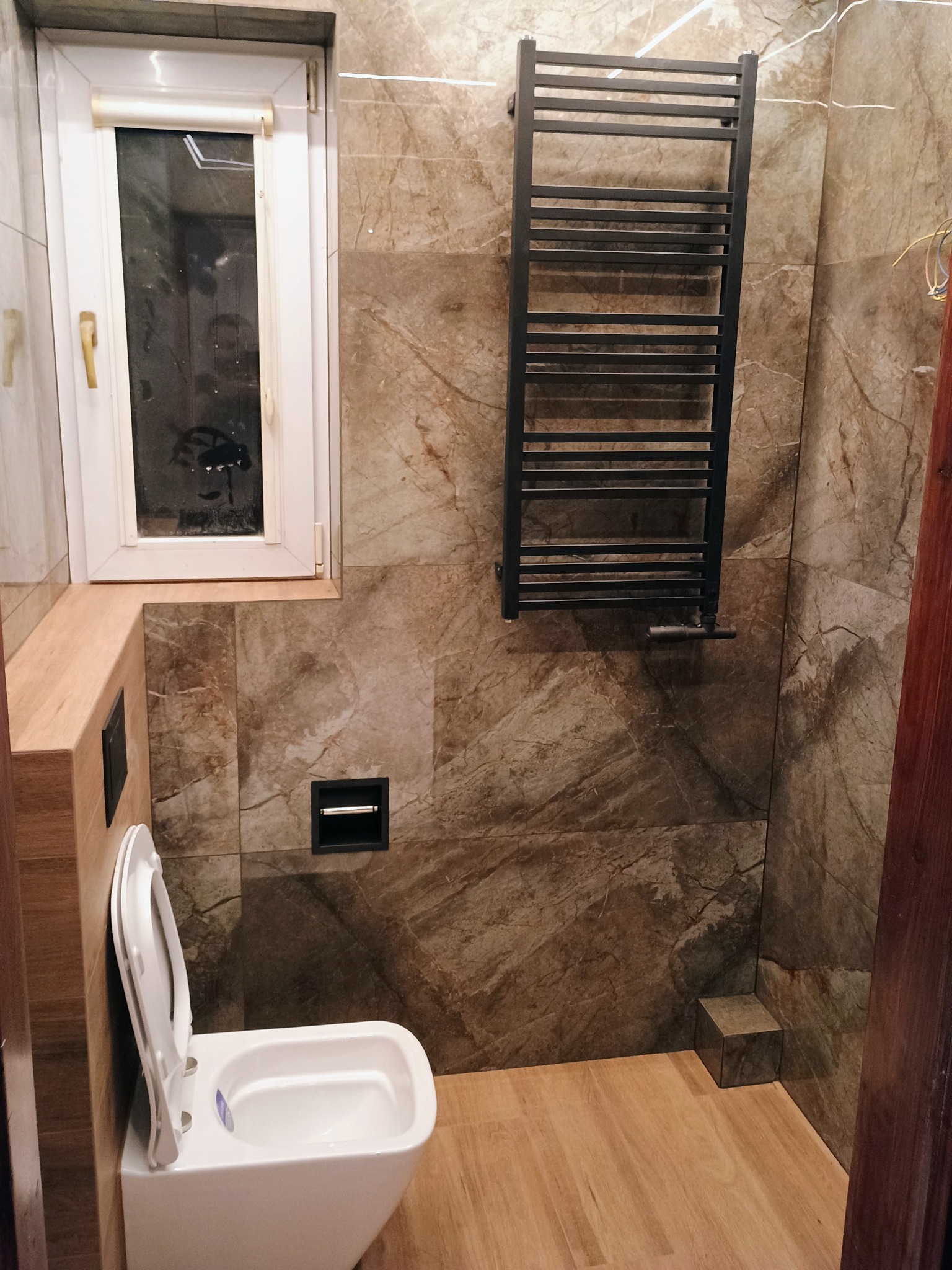 Wykończona łazienka z brązowymi marmurkowymi płytkami, białym sedesem, grzejnikiem drabinkowym i drewnianą podłogą, widoczne niedociągnięcia w postaci wystających kabli.