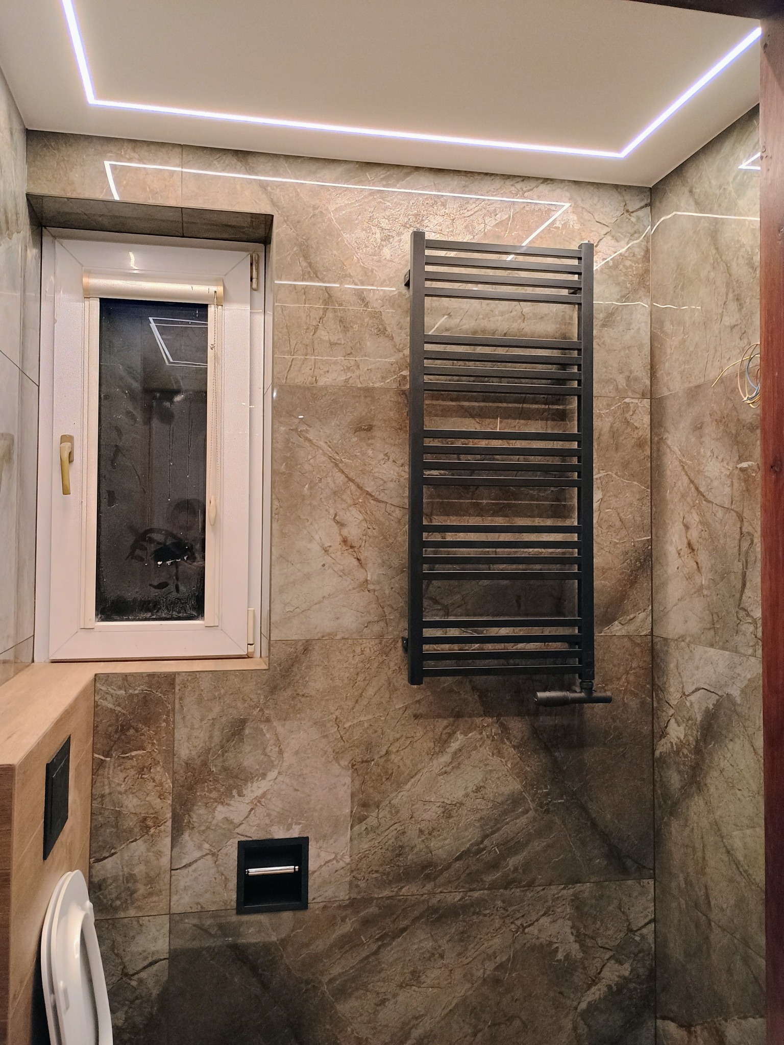 Wykończona łazienka z marmurowymi płytkami, grzejnikiem drabinkowym w kolorze antracytowym, podświetlanym sufitem LED i oknem, na którym widnieje rysunek palcem.
