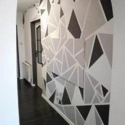 Wnętrze korytarza z dekoracyjnym malowaniem ścian w geometryczne, trójkątne wzory w odcieniach szarości i czerni. Ciemna podłoga i widoczne drzwi w tle.