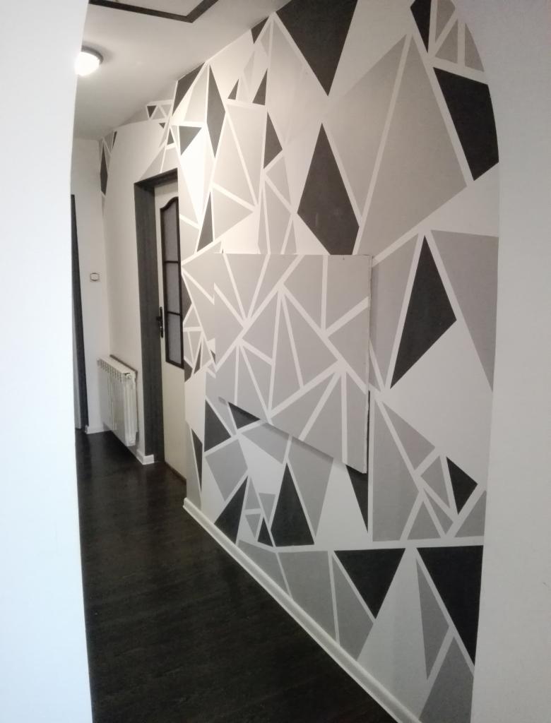 Wnętrze korytarza z dekoracyjnym malowaniem ścian w geometryczne, trójkątne wzory w odcieniach szarości i czerni. Ciemna podłoga i widoczne drzwi w tle.