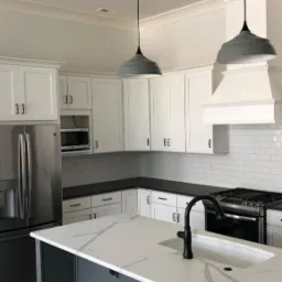 Nowoczesna, jasna kuchnia z białymi szafkami, ciemnym blatem i wyspą kuchenną w kolorze grafitowym, oświetlona wiszącymi lampami w stylu industrialnym.