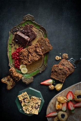 Kompozycja różnych ciast i owoców na ozdobnych talerzach, w tym brownie, ciasto owsiane, ciastka z dżemem, truskawki, miechunka i porzeczki, na ciemnym tle.