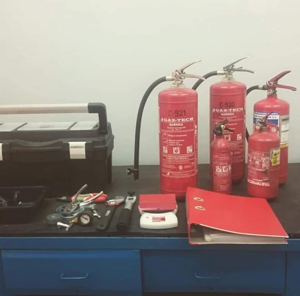 Różne gaśnice GAZ-TECH (modele E-921, E-920) stojące na warsztatowym stole obok skrzynki z narzędziami, wagi i czerwonego segregatora.