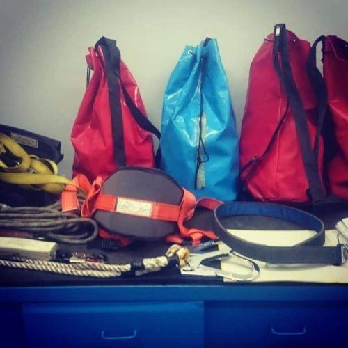 Zestaw sprzętu BHP na niebieskim biurku: czerwone i niebieskie worki transportowe, liny, uprząż asekuracyjna z etykietą, pas biodrowy i karabinki.